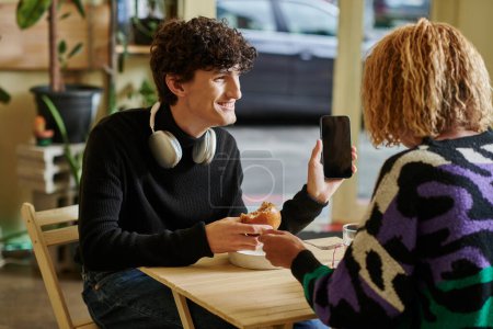 heureux bouclé homme avec casque sans fil tenant hamburger tofu et smartphone dans un café végétalien