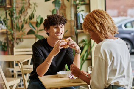 hombre contento y rizado sosteniendo hamburguesa de tofu cerca de chica afroamericana disfrutando de ensalada en la cafetería vegana