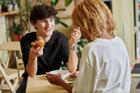 hombre sonriente y rizado comiendo hamburguesa de tofu cerca de chica afroamericana disfrutando de ensalada en la cafetería vegana