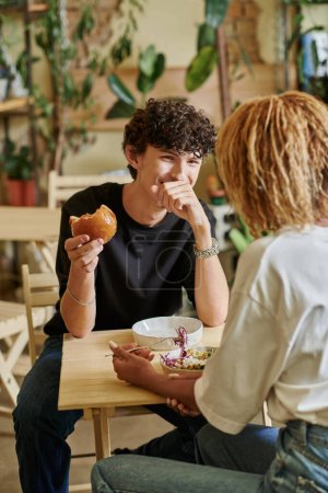 pleased curly guy eating tofu burger near african american girl enjoying salad in vegan cafe
