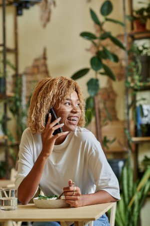 gai afro-américaine fille dans bretelles parler sur smartphone et manger de la salade végétalienne fraîche dans le café