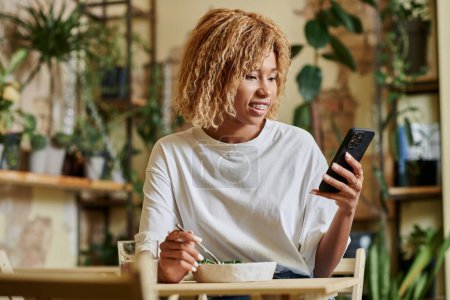 cheerful african american girl in braces using smartphone and eating fresh vegan salad in cafe