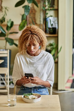 focused young african american woman using smartphone near fresh vegan salad bowl in cafe