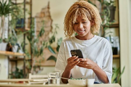 happy african american woman in braces using smartphone near fresh vegan salad bowl in cafe
