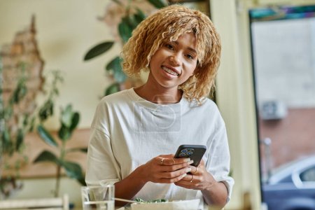 fröhliche Afroamerikanerin in Hosenträgern mit Smartphone in der Nähe einer frischen veganen Salatschüssel im Café