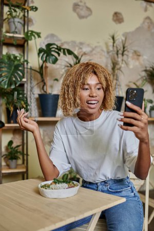 excited dark skinned young woman with braces holding fork near vegan salad and looking at smartphone