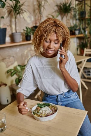 mujer joven de piel oscura feliz con frenos que sostienen tenedor cerca de ensalada vegana y hablando en el teléfono inteligente