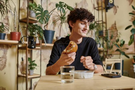 happy and young curly man holding tofu burger and looking at smartphone on table in vegan cafe