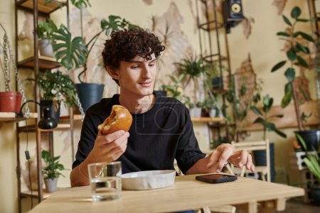 happy and young curly man holding tofu burger and using smartphone on table in vegan cafe
