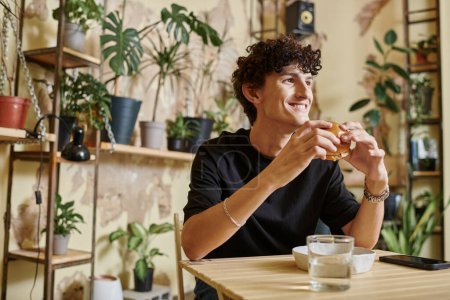 happy young man holding tofu burger and looking away while smiling in vegan cafe, enjoyment