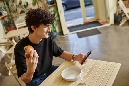 cheerful young curly man holding plant-based tofu burger and using smartphone in vegan cafe