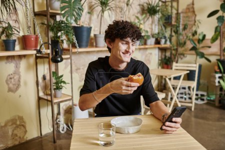 hombre rizado joven positivo sosteniendo hamburguesa de tofu a base de plantas y el uso de teléfono inteligente en la cafetería vegana