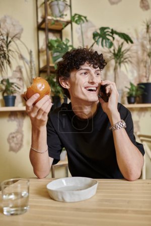 hombre rizado joven positivo sosteniendo hamburguesa de tofu a base de plantas y hablando en el teléfono inteligente en la cafetería vegana