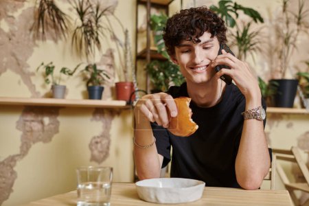 joven feliz y rizado sosteniendo hamburguesa de tofu a base de plantas y hablando en el teléfono inteligente en la cafetería vegana