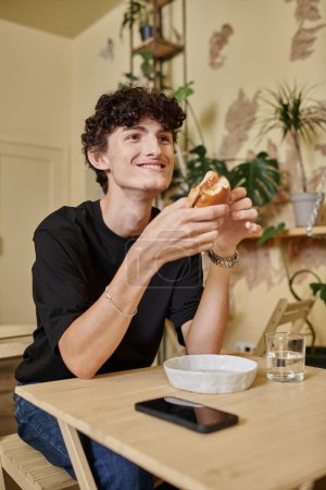 joven positivo y rizado sosteniendo hamburguesa de tofu orgánico y sonriendo en la planta llena de café vegano