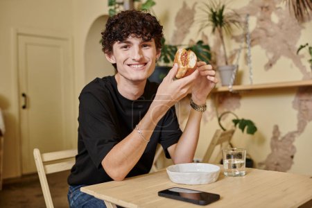 cheerful and curly young man holding organic tofu burger and smiling in plant filled vegan cafe