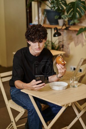 joven feliz y rizado sosteniendo a base de plantas de tofu hamburguesa mensajes de texto en el teléfono inteligente en la cafetería vegana