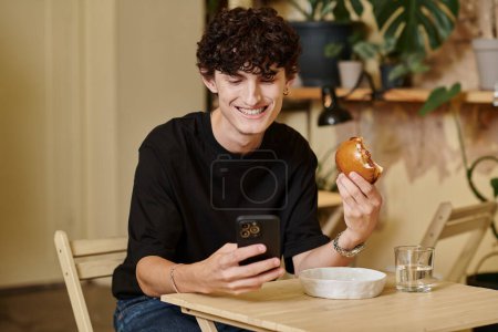 smiling and curly young man holding plant-based tofu burger texting on smartphone in vegan cafe