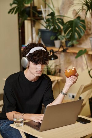 young man in wireless headphones using laptop and enjoying tofu burger in vegan cafe, plant based