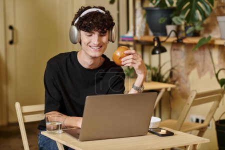cheerful man in wireless headphones using laptop and enjoying tofu burger in vegan cafe, plant based
