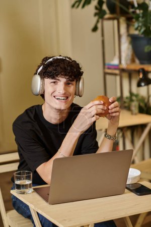 hombre positivo en auriculares inalámbricos utilizando el ordenador portátil y disfrutar de hamburguesa de tofu en la cafetería vegana, a base de plantas