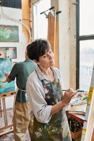 morena reflexiva mujer madura pintura en caballete cerca de amiga en taller de arte, creatividad