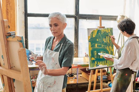 mujer madura de pelo gris en la pintura de delantal en caballete cerca de una amiga en el taller de arte, creatividad