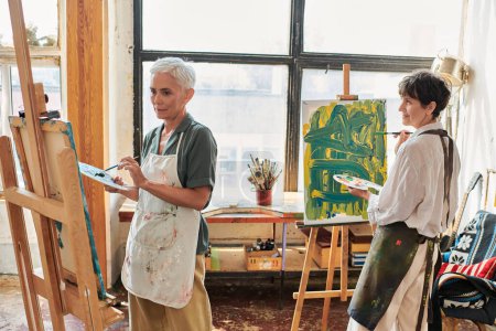 talentierte reife Freundinnen in Schürzen malen auf Staffeleien in der Werkstatt für moderne Kunst, kreatives Hobby