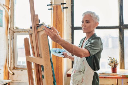 mujer madura de pelo gris concentrado en delantal mirando caballete en taller de artesanía, hobby creativo