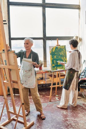talentosas mujeres de mediana edad en delantales pintura sobre caballetes en el taller de arte moderno, hobby creativo