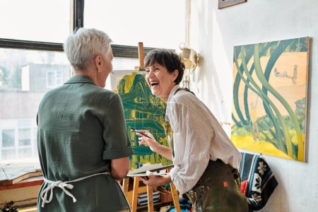 mujer madura emocionada riendo cerca del caballete y amiga en taller de artesanía, hobby creativo