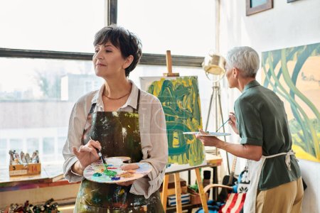 mujer madura inspirada sosteniendo paleta mientras que la pintura amiga femenina en el taller de arte, ocio creativo