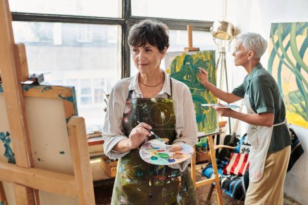 brünette reife Frau schaut auf Staffelei, während Freundin Malerei in Kunstwerkstatt, kreatives Hobby