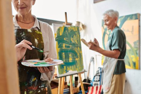 mujer madura con paleta cerca sonriente amiga pintura sobre caballete en taller de arte, creatividad