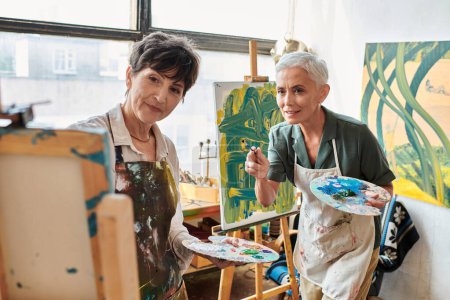 mujeres artistas maduras con paletas mirando caballete en taller de arte, creatividad y colaboración