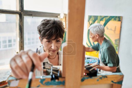 Photo for Brunette mature woman near easel and female girlfriend painting on blurred backdrop, artistic hobby - Royalty Free Image