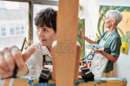 Photo for Creative mature woman near easel and female girlfriend painting on blurred backdrop, artistic hobby - Royalty Free Image