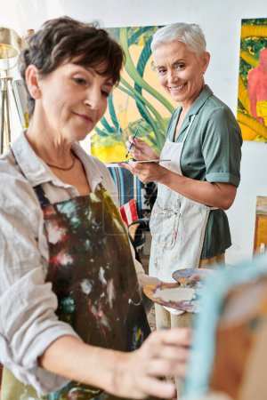 cheerful mature woman in apron looking at female friend painting on blurred foreground, creativity