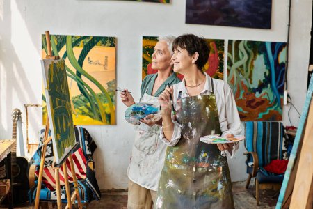 heureux femmes mûres élégantes dans des tabliers regardant loin dans l'atelier d'art moderne, l'amitié et la créativité