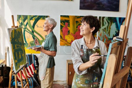sonrientes mujeres maduras en delantales pintando en caballetes y mirando hacia otro lado en el taller de arte, hobby creativo