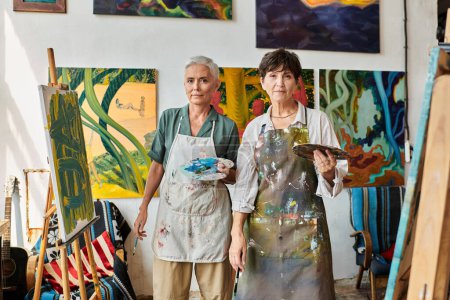 mujeres maduras expertas en delantales sosteniendo paletas y mirando a la cámara en el estudio de arte, hobby creativo