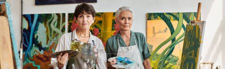 talentosas mujeres pintoras en delantales sosteniendo paletas y mirando a la cámara en el estudio de arte, pancarta