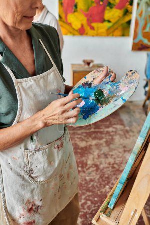 cropped view of mature female artist mixing on colors on palette with brush in art workshop