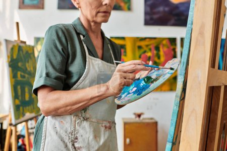 vista parcial de mujer madura en delantal con paleta y pintura de pincel sobre caballete en estudio de arte