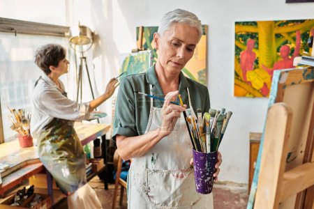 mujer madura en delantal elegir el pincel cerca de la pintura amiga en el estudio de arte, hobby creativo