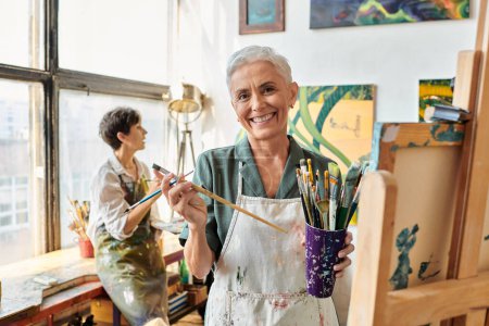 Foto de Mujer madura alegre con conjunto de pinceles cerca de amiga en taller de arte, pasatiempo creativo - Imagen libre de derechos