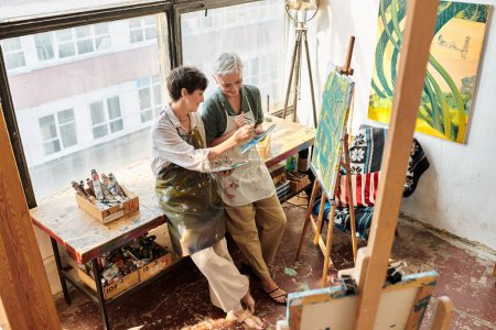 glückliche Frauen mittleren Alters beim Mischen von Farben auf Paletten in der Nähe von Staffeleien im Kunstatelier, kreatives Hobby