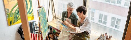 sonrientes mujeres de mediana edad mezclando colores en paletas cerca de caballetes en el estudio de arte, pancarta horizontal