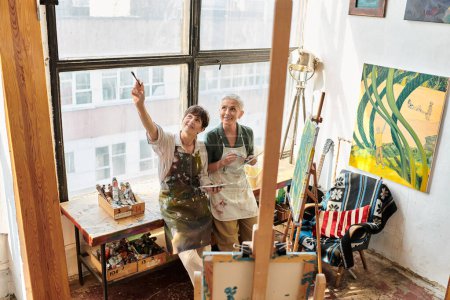 cheerful mature woman pointing up with paintbrush near female friend in art workshop, creative hobby