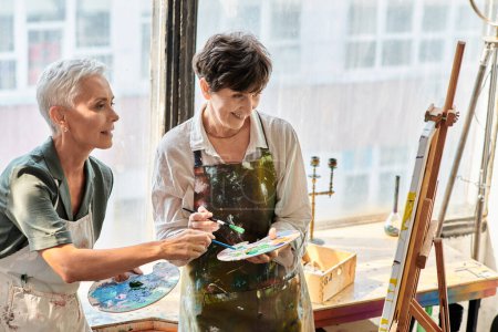 mature female artists mixing colors on palettes near easel in craft workshop, painting master class